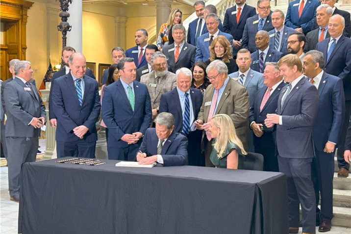 A large group of people gather around a man and a woman as the man signs a piece of paper. They are in a large atrium of a state capitol building.