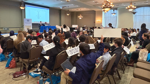 Middle school boys and girls sit in chairs facing the front of a large conference room. At the front of the room sit two college students at a Junior Model United Nations table. The children in the room have signs that read different countries' names. One boy holds his sign up.