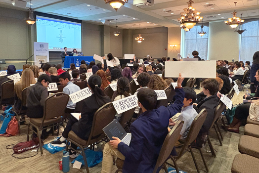 Middle school boys and girls sit in chairs facing the front of a large conference room. At the front of the room sit two college students at a Junior Model United Nations table. The children in the room have signs that read different countries' names. One boy holds his sign up.