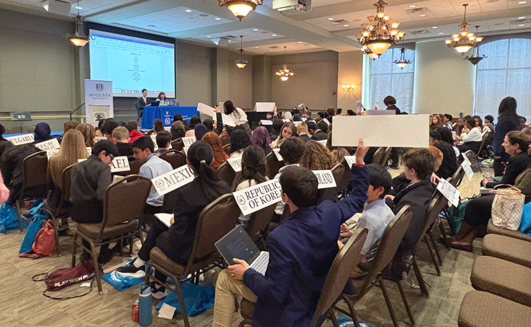 Middle school boys and girls sit in chairs facing the front of a large conference room. At the front of the room sit two college students at a Junior Model United Nations table. The children in the room have signs that read different countries' names. One boy holds his sign up.