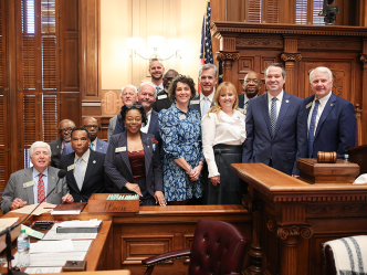 Small group of men and women pose in a chamber with a gavel visible and an American flag behind them