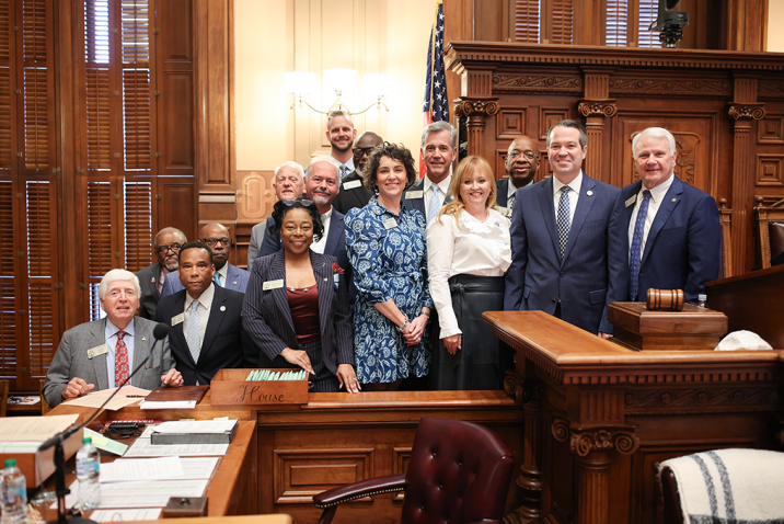 Small group of men and women pose in a chamber with a gavel visible and an American flag behind them