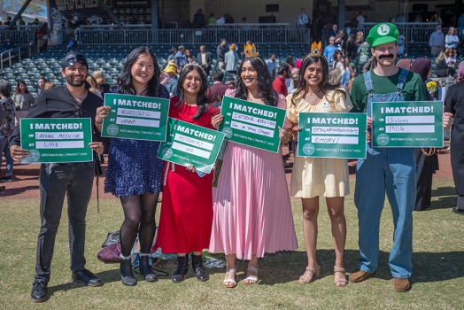 4 young women and 2 young men dressed in costume smile and hold up a small sign.