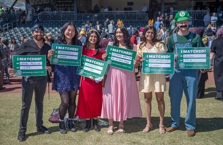 4 young women and 2 young men dressed in costume smile and hold up a small sign.