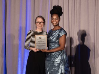 Two women stand on a stage during an awards ceremony. The woman on the right is handing the other a plaque.