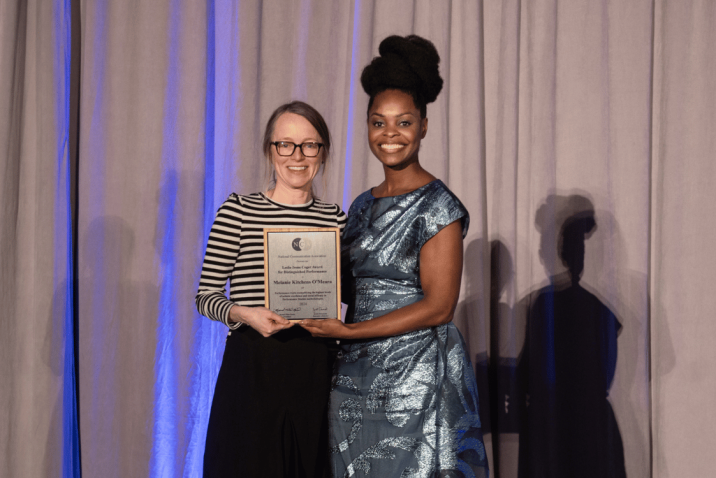 Two women stand on a stage during an awards ceremony. The woman on the right is handing the other a plaque.