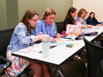 Six women sit at a long table and work on a project that includes cutting out phrases and restructuring them.