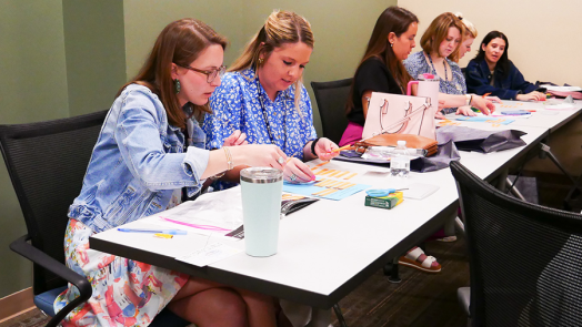 Six women sit at a long table and work on a project that includes cutting out phrases and restructuring them.
