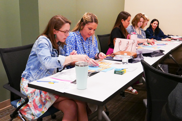 Six women sit at a long table and work on a project that includes cutting out phrases and restructuring them.