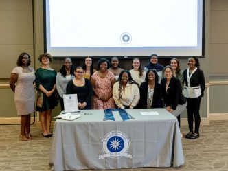 Female first-generation scholars stand gathered in front of an Alpha Alpha Alpha table.