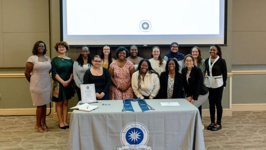 Female first-generation scholars stand gathered in front of an Alpha Alpha Alpha table.