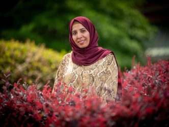 Smiling woman in a hijab and patterned blouse standing outdoors among red and green bushes.