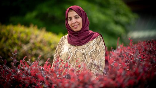 Smiling woman in a hijab and patterned blouse standing outdoors among red and green bushes.
