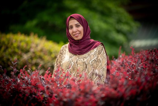 Smiling woman in a hijab and patterned blouse standing outdoors among red and green bushes.
