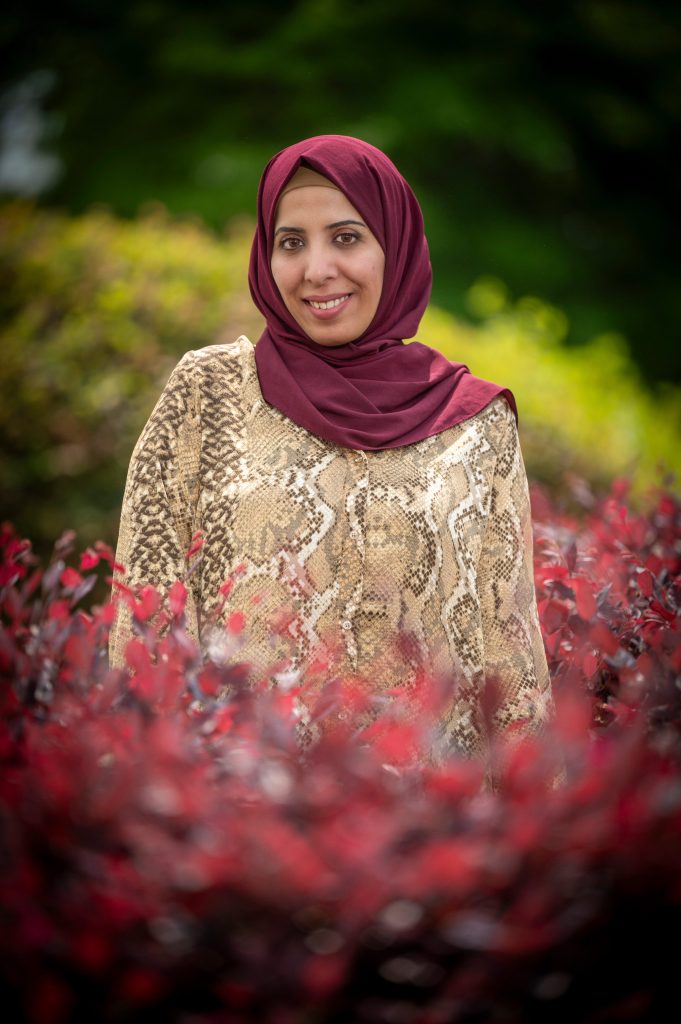 A woman wearing a hijab is posing in a photograph surrounded by red and green bushes.
