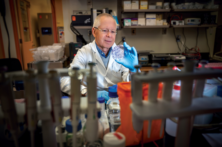 A scientist wearing a lab coat, safety glasses and gloves holds up a petri dish and examines it. He is sitting in a scientific lab.