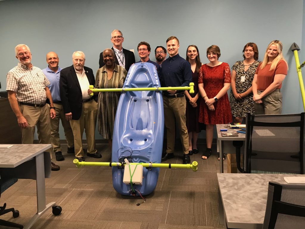 A group of people in business casual dress stand behind a blue kayak with yellow PVC rods on both vertical ends of the kayak. There is an electronic box on the back of the kayak.