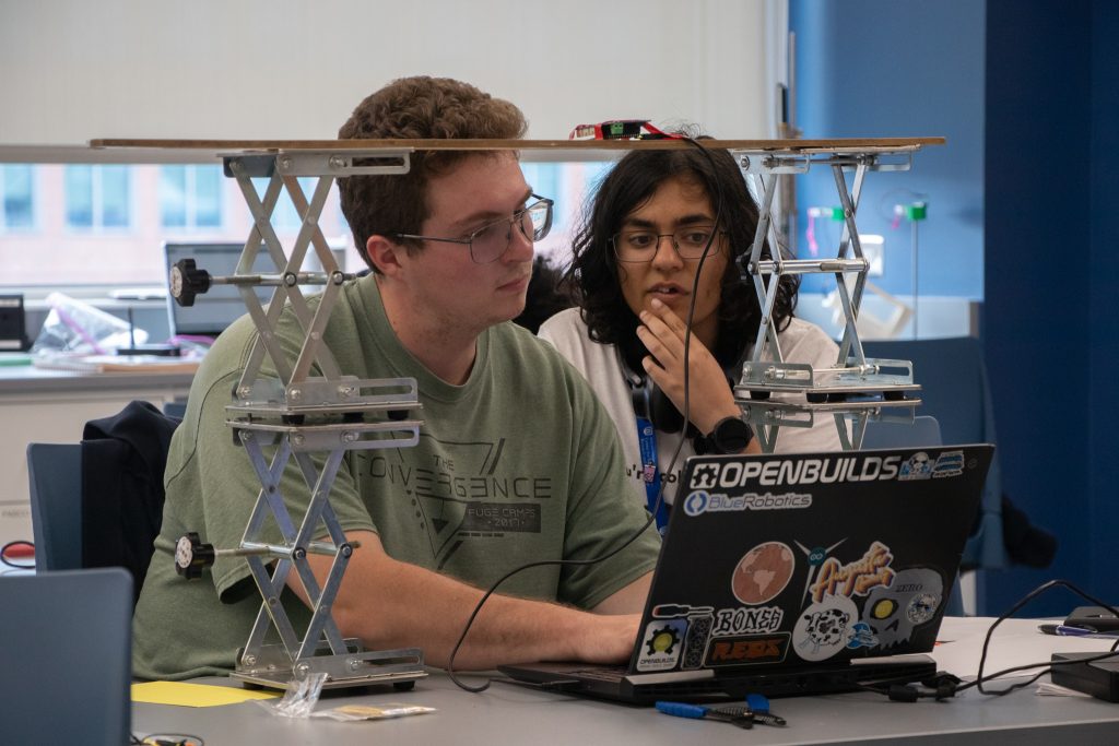 A student with brown hair, glasses, and a green shirt types on a laptop while a student with long black hair, glasses, and a white shirt looks at the laptop screen. They are surrounded by wires and electronics equipment.