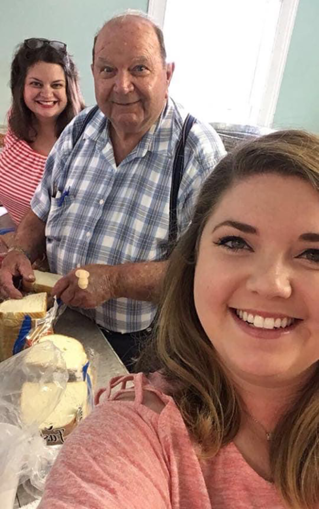 Two smiling women and a man take a selfie. The man is making a sandwich.