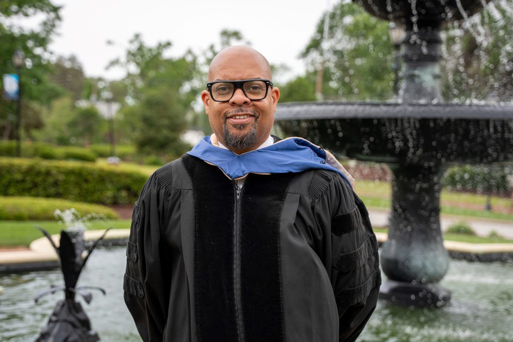 A man in a graduation gown stands in front of a fountain.