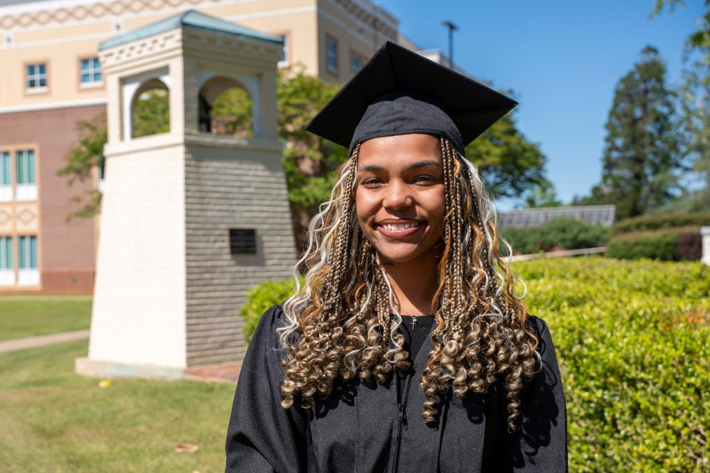 A girl in a graduation cap and gown smiles outside.