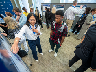 A female college student talks about a research project to another college student. They are in a large ballroom surrounded by other faculty and students.
