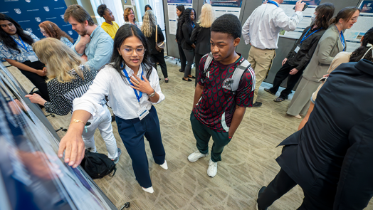A female college student talks about a research project to another college student. They are in a large ballroom surrounded by other faculty and students.