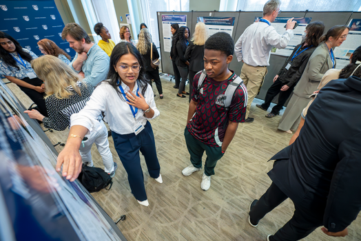 A female college student talks about a research project to another college student. They are in a large ballroom surrounded by other faculty and students.