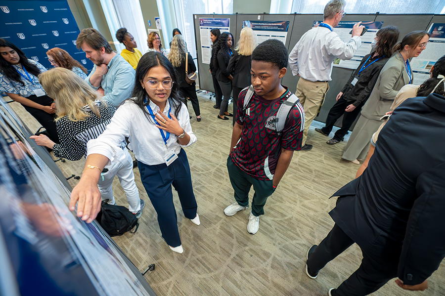 A female college student talks about a research project to another college student. They are in a large ballroom surrounded by other faculty and students.