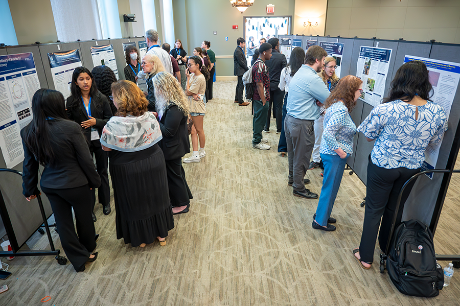 A group of college students talk to faculty and visitors about their research. Everyone is clumped in groups around different scientific posters in a large ballroom.