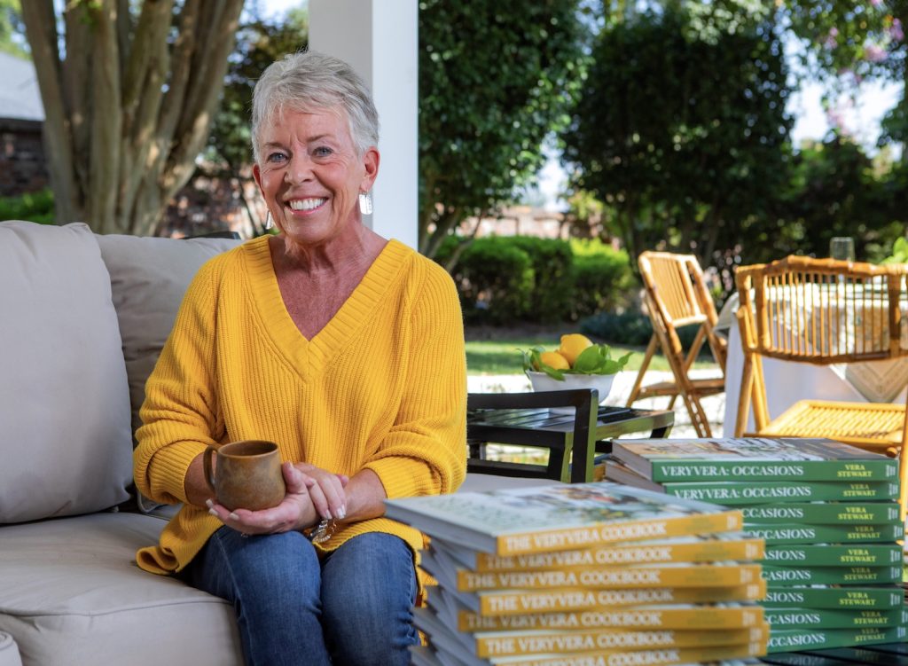 Woman sitting on a couch outside with a coffee cup in her hand and a two piles of her cookbooks in front of her on a table.