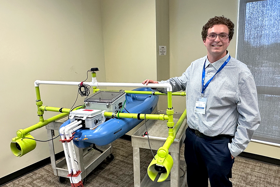 A male college student stands next to an autonomous boat he made using a child's sit-a-top kayak and PVC pipe for water testing.