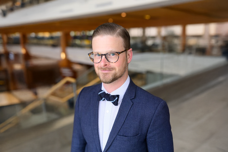 A man in a suit stands at the top of a staircase in a large, modern building on a college campus.
