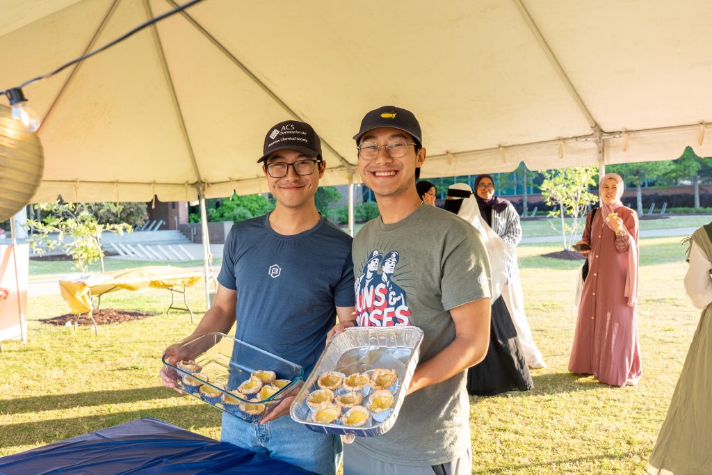 Two men standing together, both holding plates of food.