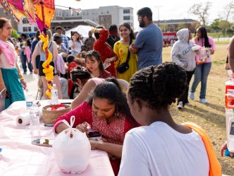 Male and female college students at an outdoor event, with some sitting at an interactive booth that provides henna designs.