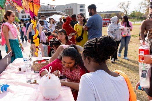 Male and female college students at an outdoor event, with some sitting at an interactive booth that provides henna designs.