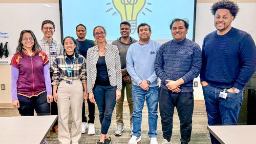 A group of college students and a professor standing in front of display with graphic of lightbulb
