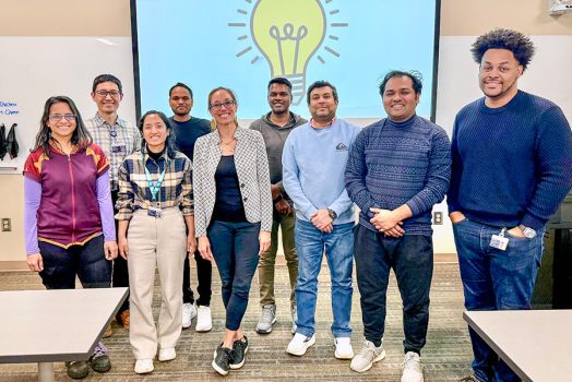A group of college students and a professor standing in front of display with graphic of lightbulb