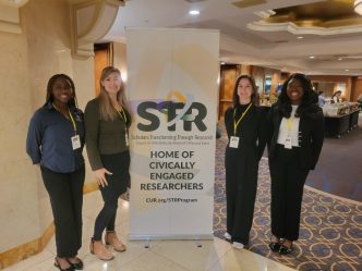 Four women standing next to a stand up poster.