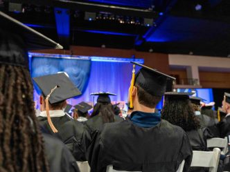 Graduating students in caps and gowns sit in an indoor event.