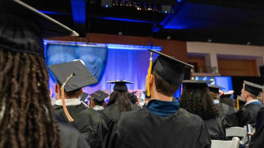 Graduating students in caps and gowns sit in an indoor event.