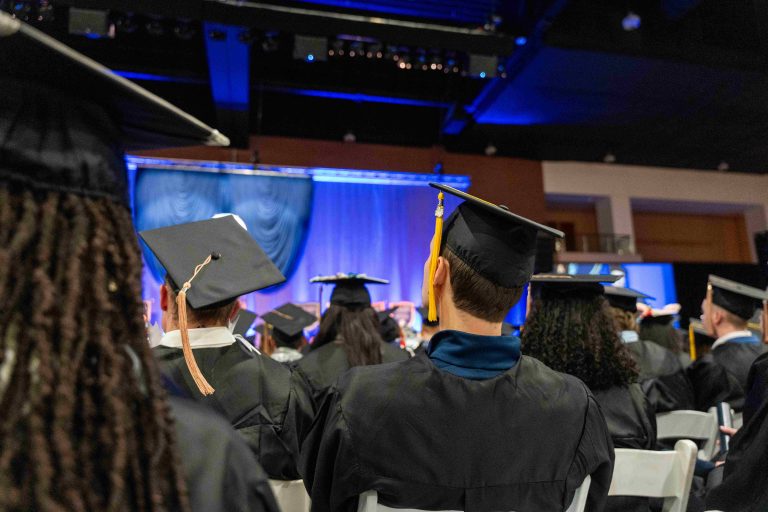 Graduating students in caps and gowns sit in an indoor event.