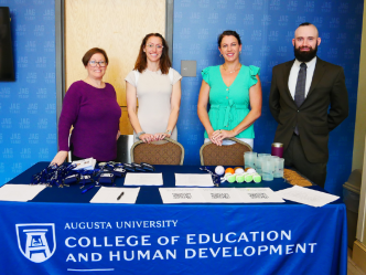 Four people, three women and one man, stand behind a table with a table cloth featuring the Augusta University College of Education and Human Development logo.