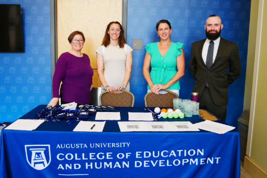 Four people, three women and one man, stand behind a table with a table cloth featuring the Augusta University College of Education and Human Development logo.