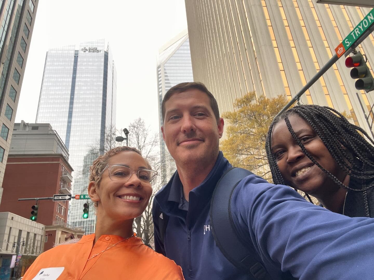 Two women and a man take a selfie in an intersection in downtown Charlotte surrounded by tall skyscraper buildings