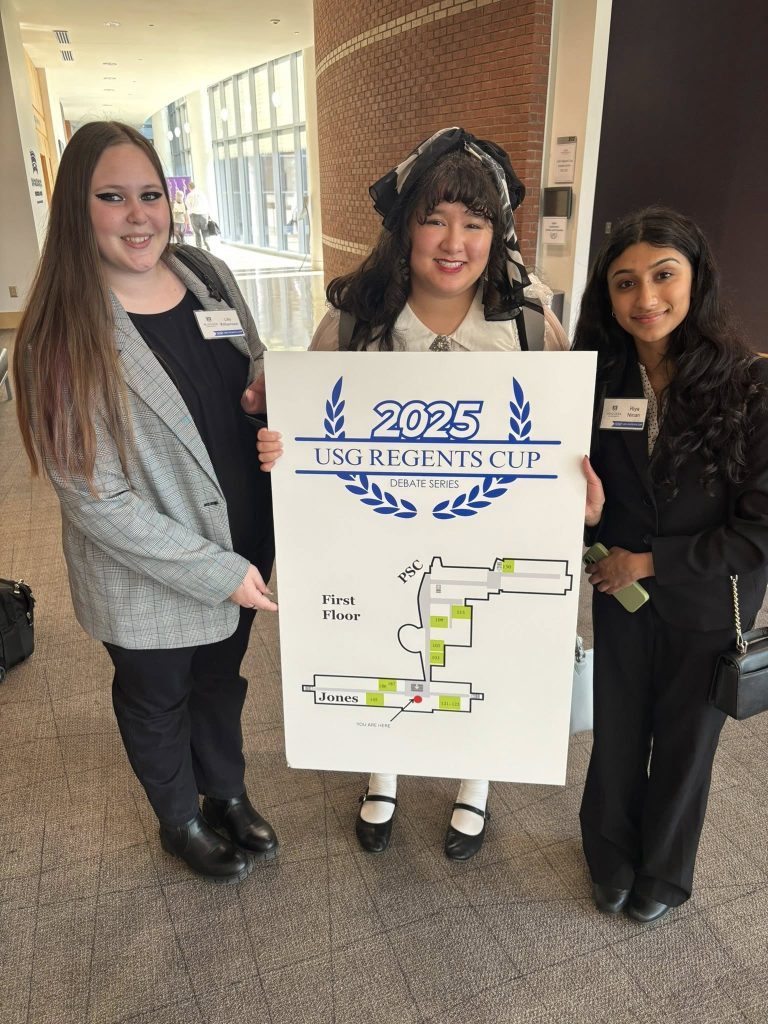 Three college students hold up a sign for the 2025 University System of Georgia Regents' Cup debate competition in a hallway.