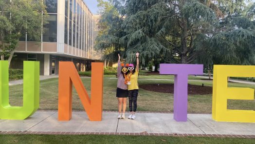 Two women form a capital I between the letters UN and TE to form "Unite" at an outdoor event.