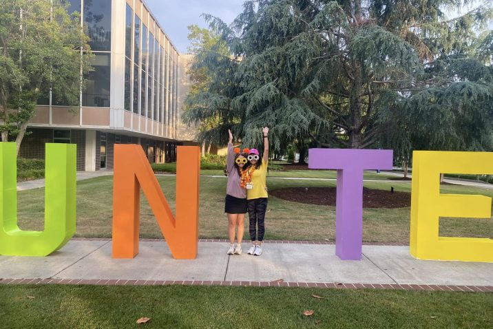 Two women form a capital I between the letters UN and TE to form "Unite" at an outdoor event.