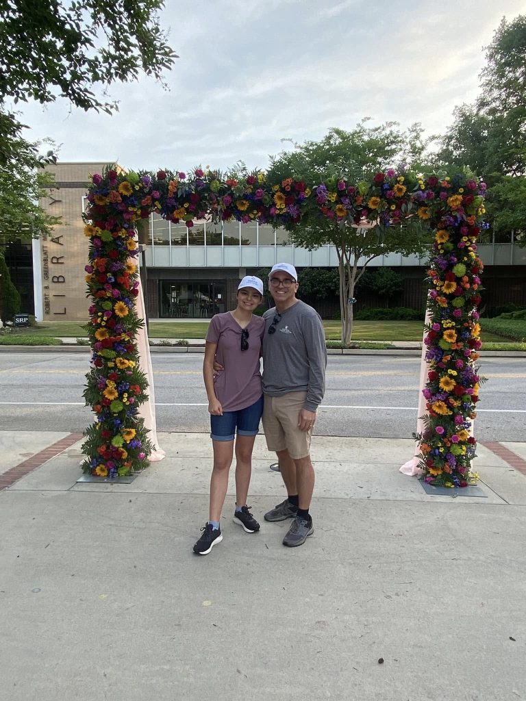 A man and a woman stand outside under an arch made out of flowers.