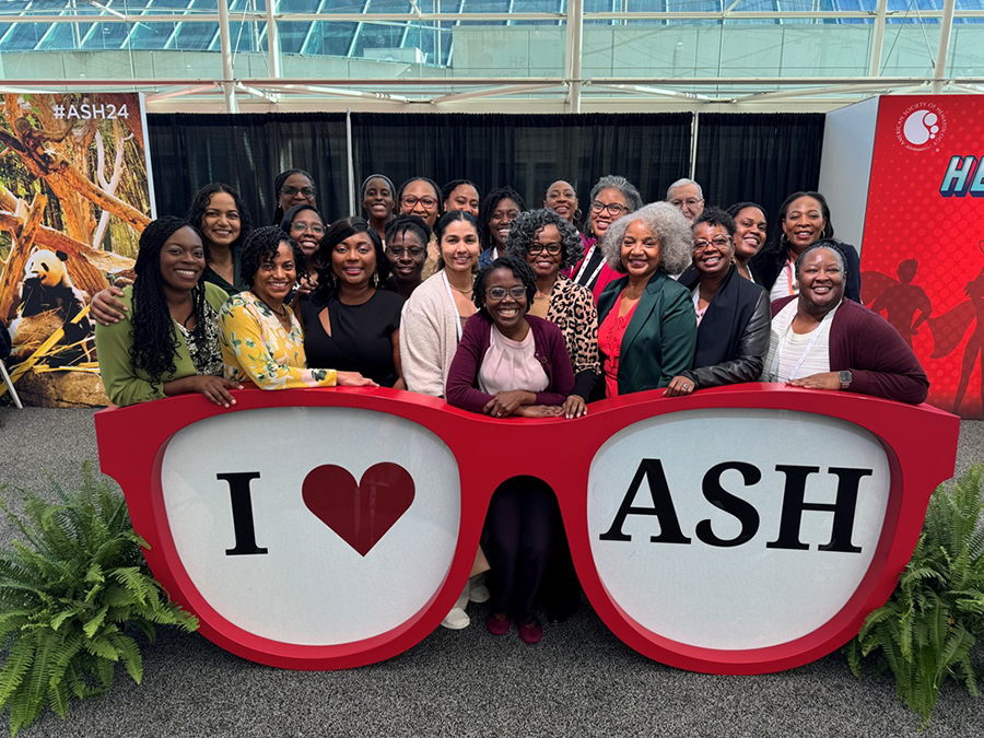A group of women stand behind an over-sized pair of sunglasses with the words "I love ASH" written on them. ASH stands for American Society of Hematology.